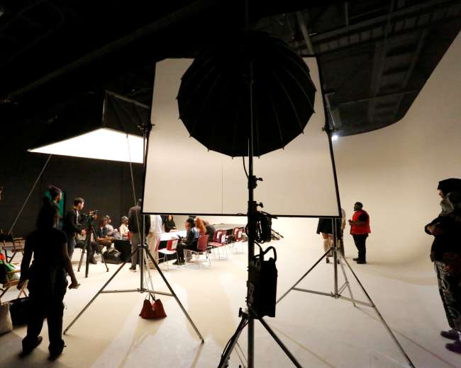A film studio setup features large lighting equipment and a backdrop with crew members and models preparing for a shoot.