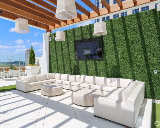 The image shows a modern outdoor lounge area with a large white sectional sofa, round seating, and a wall of greenery under a pergola with hanging light fixtures and a television.
