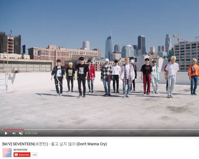 A group of twelve performers stands on a rooftop with a city skyline in the background.