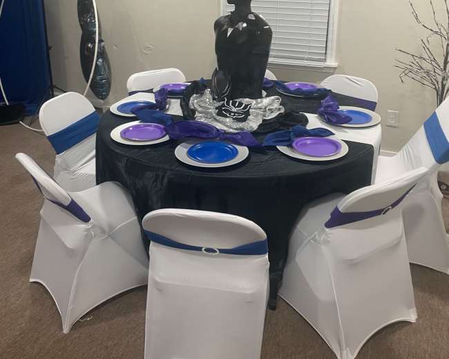 A round table with black and purple decorations is set with plates and a central black statue surrounded by six chairs.