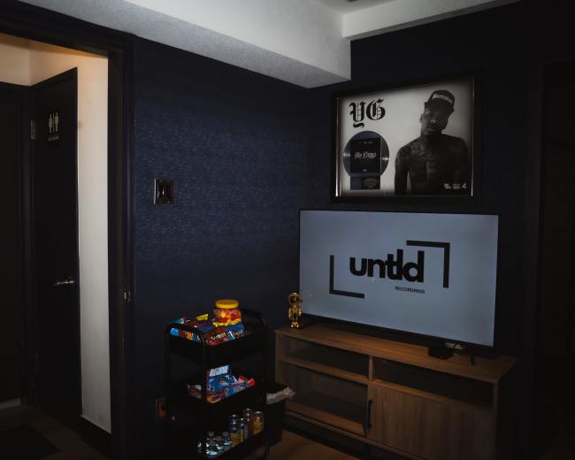 The image shows a dimly lit room featuring a television with a logo for "untld recordings," a framed poster on the wall, and shelves with various snacks and a small trophy.