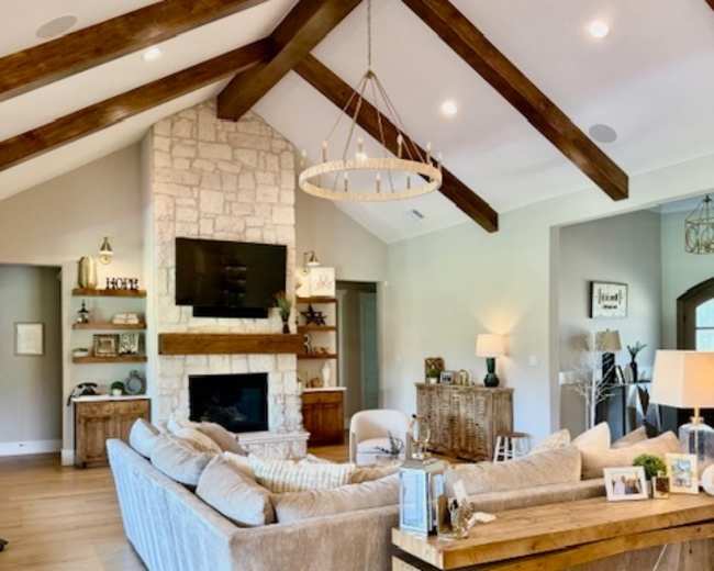 The image shows a spacious living room featuring a large beige sectional sofa, a stone fireplace with a television above it, and exposed wooden beams on the ceiling.