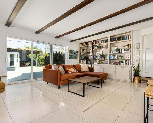 A modern living room with a brown sectional sofa, a coffee table, built-in shelves filled with books and decor, and large windows overlooking an outdoor patio.