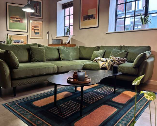 The image features a modern living room with a green sectional sofa, a round coffee table on a patterned rug, and framed art on the walls.