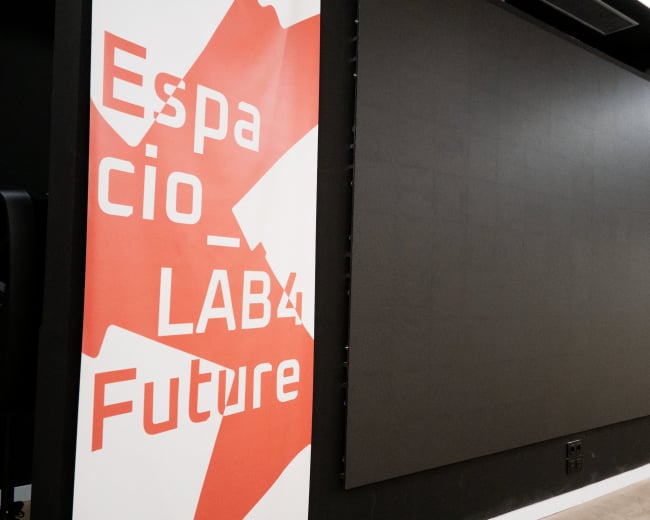 The image shows a large blank screen mounted on a wall, flanked by banners with text on a red background promoting "Espacio LAB4 Future."