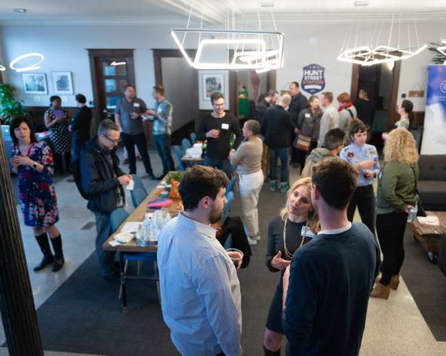 A group of people socializes at a networking event in a modern indoor space with a lounge area and refreshments laid out on a table.