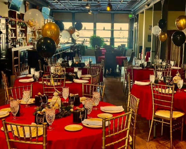 The image shows a elegantly arranged dining room with red tablecloths, black and gold balloons, and festive decorations, set for a special event.