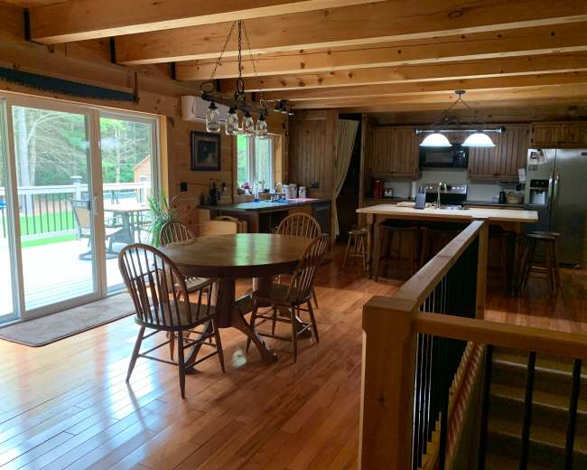 The image shows a wooden cabin interior featuring a dining area with a round table and four chairs, a kitchen with modern appliances, and large sliding glass doors leading to a deck.