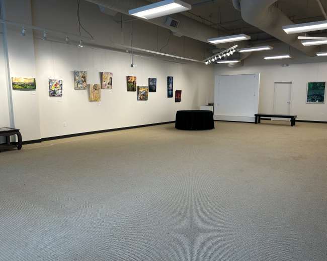 The image shows a spacious art gallery with several paintings displayed on the walls and a round table in the center.