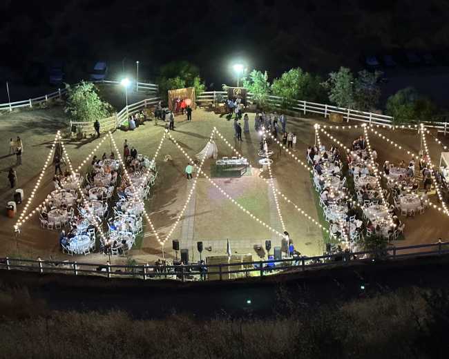 An outdoor event space is set up with multiple round tables decorated with white tablecloths, surrounded by string lights and greenery, while attendees gather in a festive atmosphere.