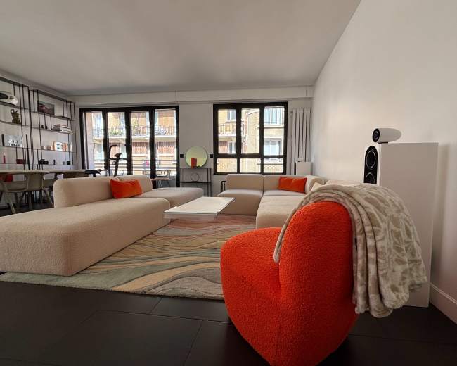 A modern living room features a light-colored sofa, an orange accent chair, a white coffee table, and large windows overlooking a cityscape.