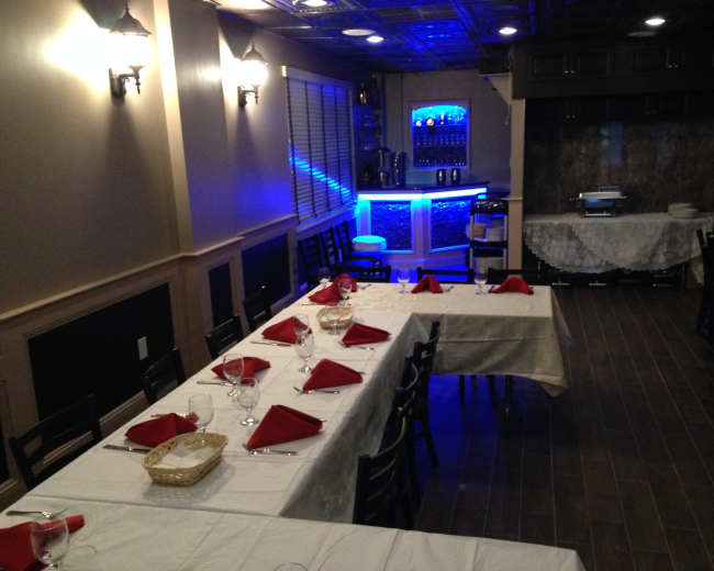 A restaurant dining area with neatly arranged tables covered in white tablecloths and red napkins, featuring a bar area illuminated by blue lighting.