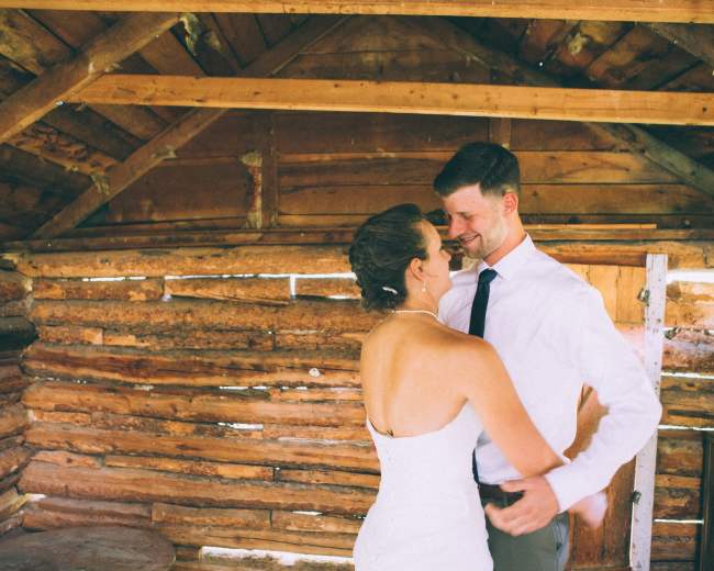 A couple stands together inside a rustic cabin with wooden walls and a slanted roof.