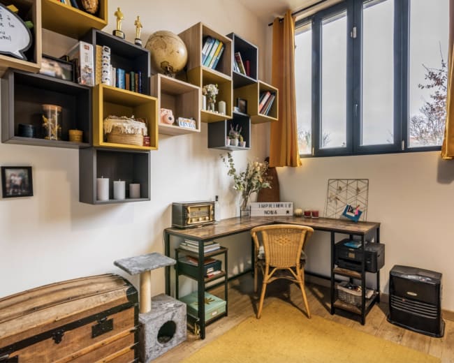 A cozy office space features a desk with a wicker chair, shelves filled with books and decor, and a large window with yellow curtains.