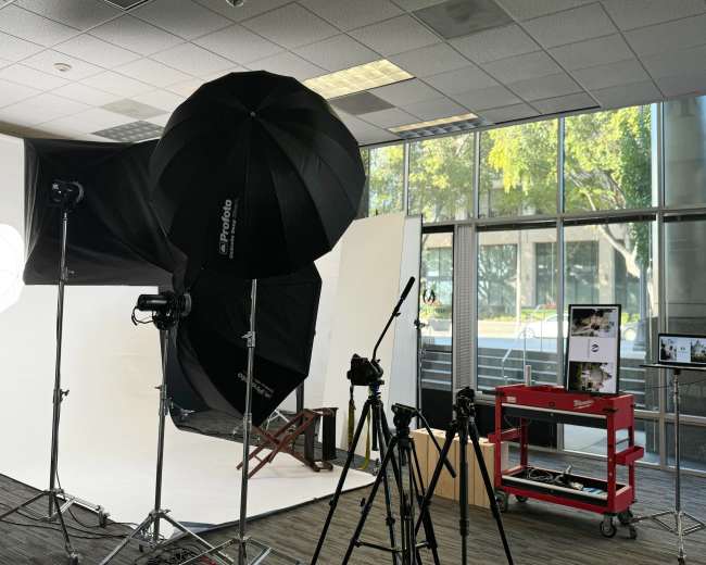 A photography studio setup features softbox lights, tripods, and a red cart with equipment, all positioned in front of a large window with visible greenery outside.