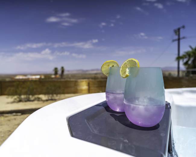 Two glasses with purple drinks and lemon slices are positioned on the edge of a hot tub, with a desert landscape visible in the background.