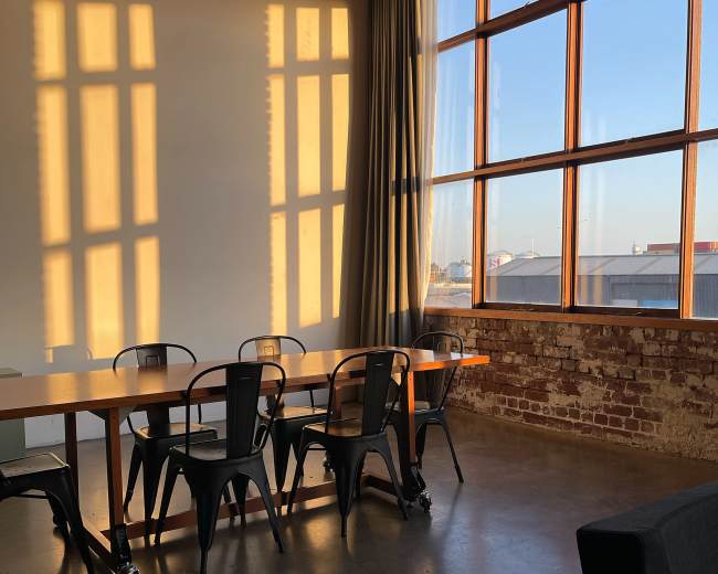 A long wooden table with metal chairs is positioned near large windows, casting shadows on the wall as sunlight streams into the room.