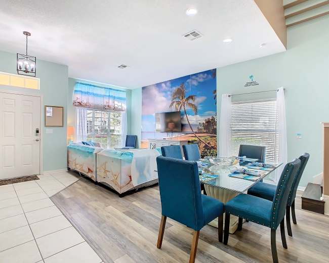 The image shows a bright dining area with a glass table set for a meal, surrounded by blue upholstered chairs, and a large wall mural depicting a beach scene.