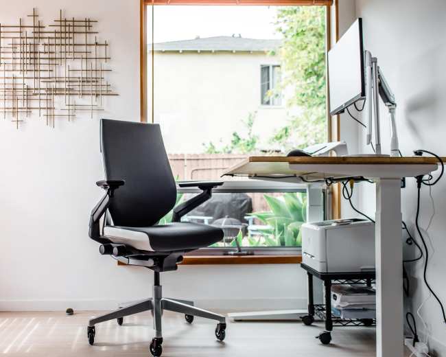 A black ergonomic chair is positioned in front of a desk with a computer monitor, beside a large window with wooden blinds.