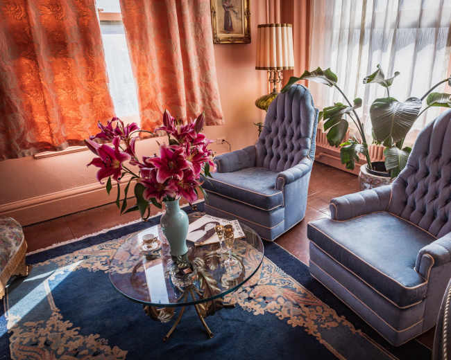 The image shows a cozy living room with two blue upholstered chairs, a glass coffee table with a floral arrangement, and large windows draped with orange curtains.