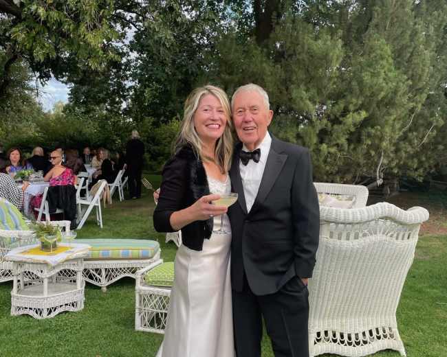 A woman in a formal dress and a man in a tuxedo stand together in a garden setting with guests seated in the background.