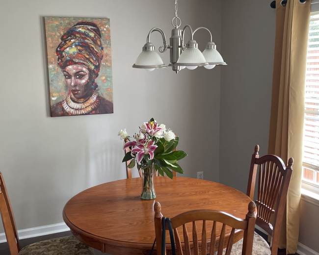 A round wooden dining table is set in a room with a colorful painting on the wall, a vase of flowers in the center, and a light fixture hanging above.