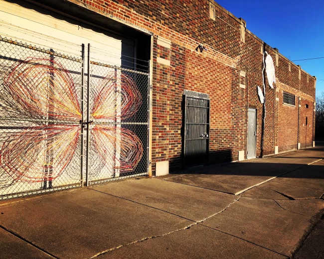 A brick building features a chain-link fence with a colorful mural resembling a flower on one side and a series of doors on the other.