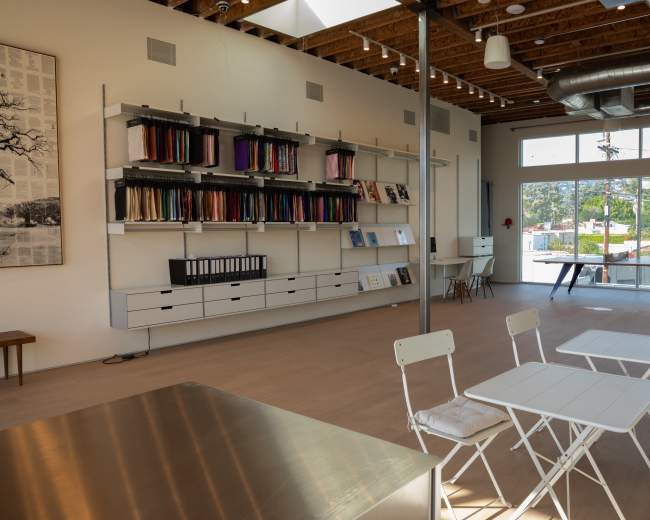 The interior of a modern workspace features a minimalist design with shelves of colorful fabrics, a long table, and seating arrangements.