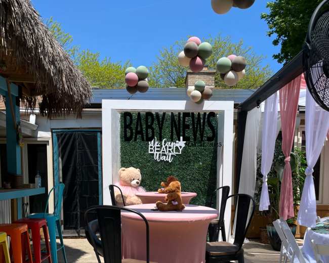 The image shows a decorated outdoor space with a circular table draped in pink, featuring two stuffed animals on top, surrounded by black bar stools and colorful decor overhead.