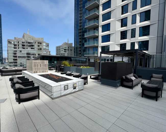 The image shows a modern rooftop terrace featuring seating areas, a fire pit, and surrounding high-rise buildings.