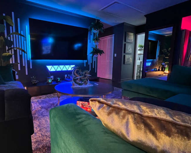 The room features a modern design with green and blue lighting, a large wall-mounted TV, and plush seating arranged around a decorative coffee table.