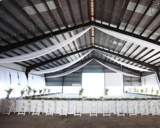 The image shows a large indoor venue with a long table set for an event, featuring white tablecloths and floral arrangements along the center, beneath a high, draped ceiling with string lights.