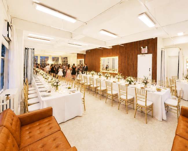 The image shows a spacious event venue set up for a formal dinner, featuring long tables with white tablecloths, gold chairs, and floral centerpieces, while guests mingle in the background.