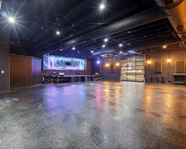 The image shows an empty performance venue with a polished floor, dim lighting, and a backdrop featuring illuminated graphics.