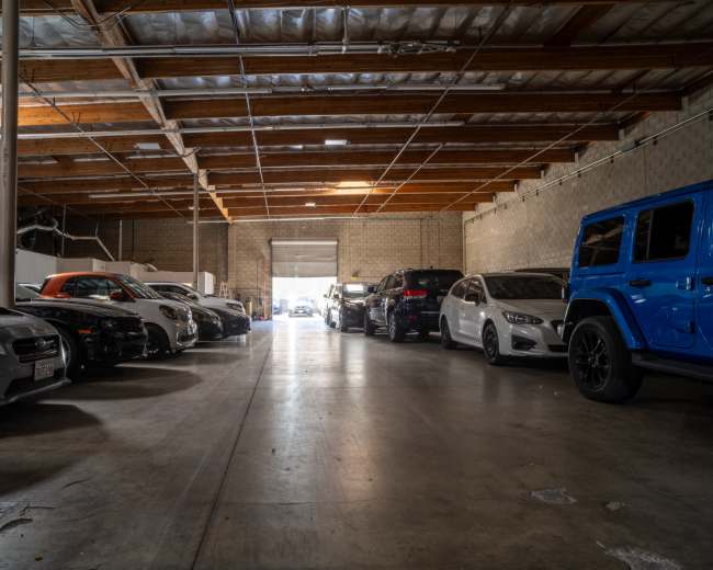 The image shows a warehouse interior with multiple parked cars lined up along both sides of a central pathway leading to an open exit at the far end.