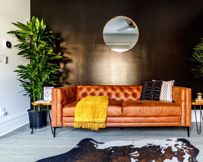 A brown leather sofa is positioned against a black wall, flanked by tall green plants, with a round mirror above and small side tables next to the sofa, one adorned with a yellow throw blanket.