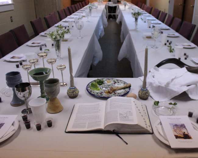 A long dining table is set with white tablecloths, glassware, and a variety of ceremonial items, including a large open book and small cups arranged along the center.