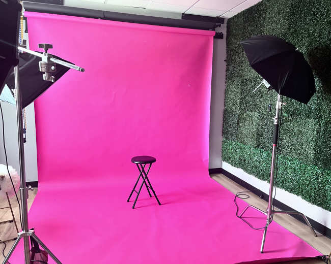 The image shows a photography studio setup with a pink backdrop, a black stool in the center, and two lighting umbrellas positioned on either side.