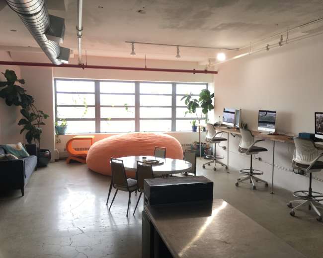 The image shows a modern, open office space featuring a large orange bean bag, a round dining table, and several workstations with computers, all illuminated by natural light from large windows.