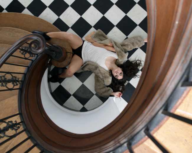 A person lies on a black and white checkered floor at the base of a wooden staircase, surrounded by a spiral railing.