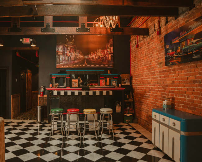 The image shows a vintage-style diner with a checkered floor, colorful stools, exposed brick walls, and a large framed photograph on the wall.