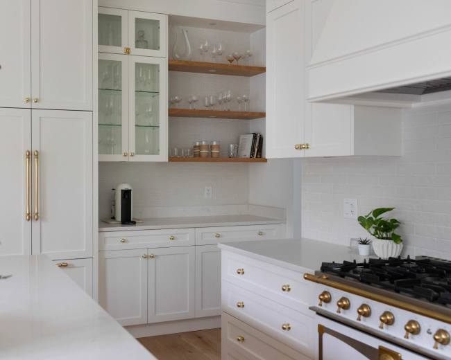 The image shows a modern kitchen with white cabinetry, glass shelves displaying glassware, and a stove with a gold-accented faucet.
