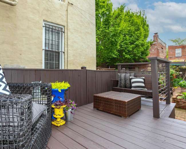 The image shows a wooden deck furnished with black wicker seating and colorful potted plants, surrounded by greenery and brick walls.