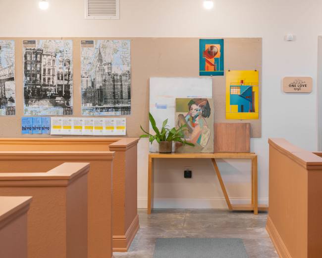 The image shows a well-lit interior of a space with wooden benches, a table displaying artwork and a plant, and walls featuring framed art and a bulletin board.