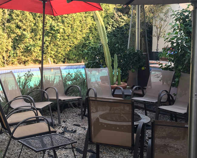 A patio area with several metal and fabric chairs arranged around a table, surrounded by greenery and a swimming pool in the background.