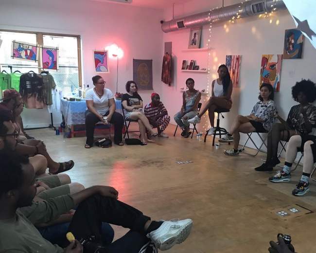 A group of people sits in a small, art-filled room, engaged in discussion while some participants are on chairs and others on the floor.