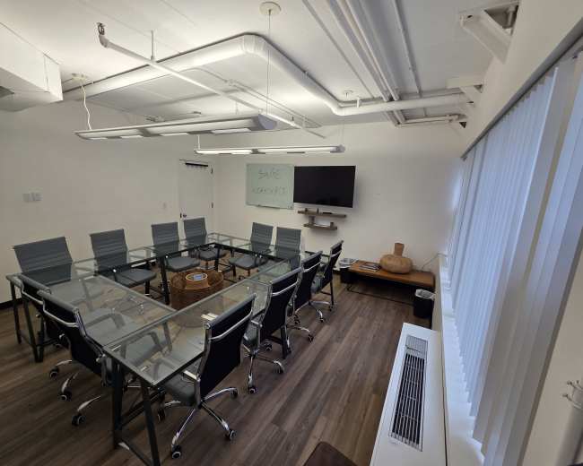The image shows a modern conference room featuring a large glass table surrounded by eight office chairs, a flat-screen TV on the wall, and a large window with vertical blinds.