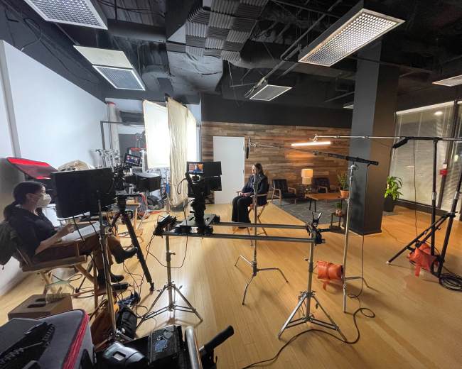 The image shows a film set with cameras and lighting equipment arranged around a woman seated in front of a backdrop.