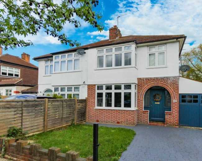 The image shows a two-story, semi-detached house with a brick and white exterior, a blue garage door, and a front garden featuring a grassy area and a wooden fence.