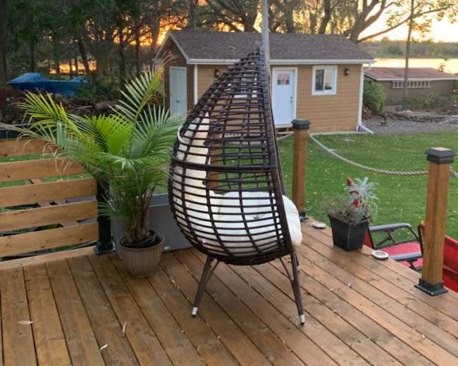 A hanging chair is placed on a wooden deck overlooking a sunset and a small cabin in the background.
